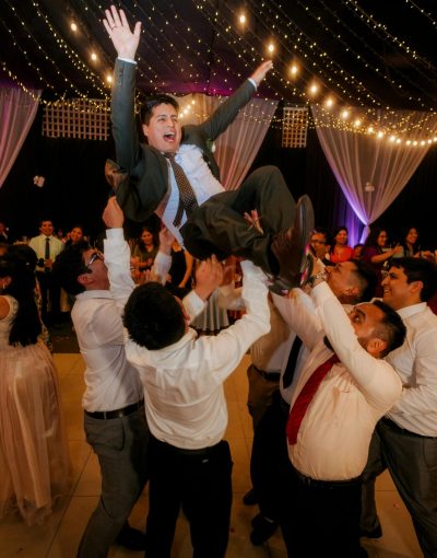 Guests celebrate joyfully at a wedding, lifting the groom amid laughter and dancing.